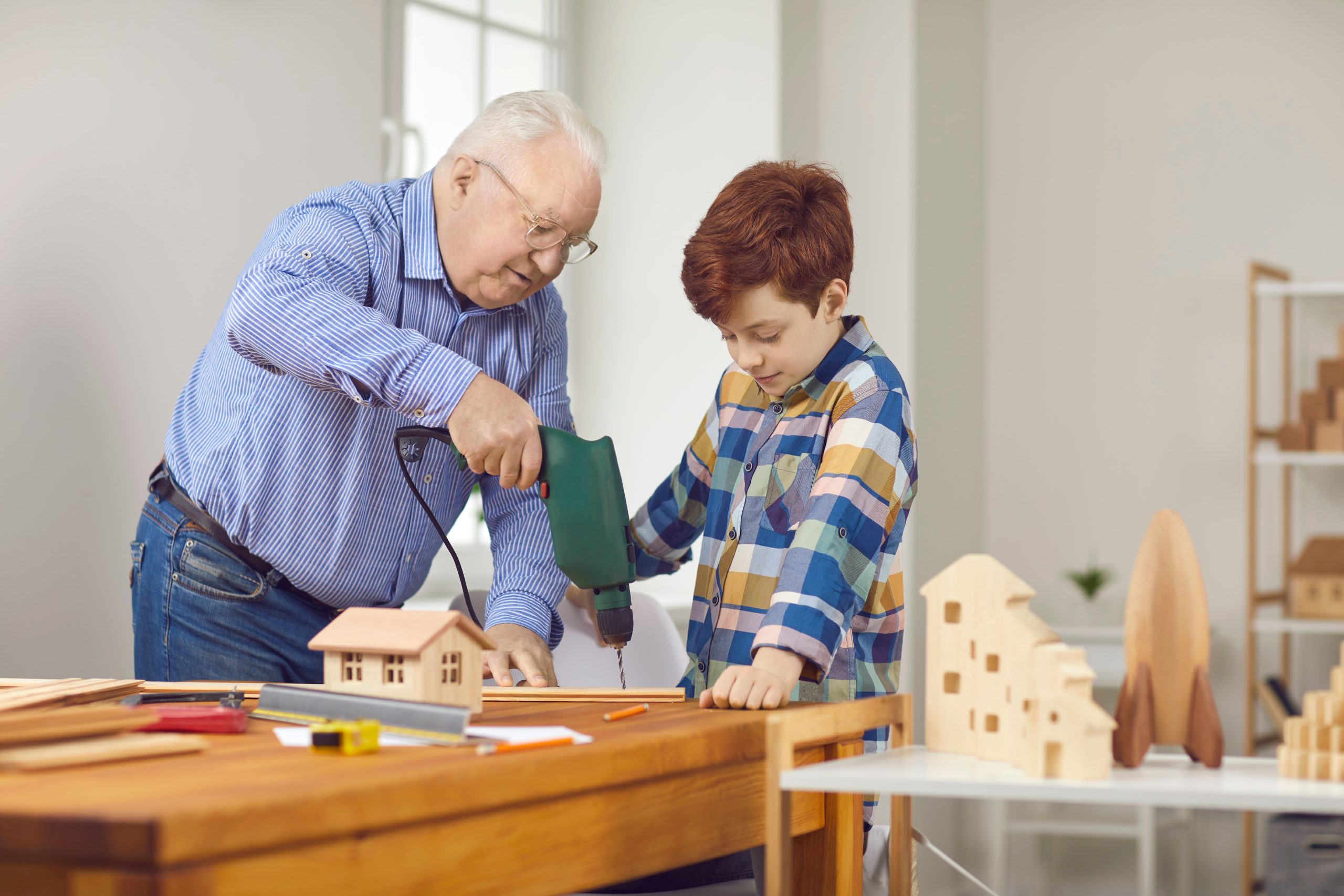 Grandfather and teen grandson working with wood and building toy houses together. Senior man teaches little child to do home repairs and explains how to use drill and other tools in carpentry workshop
